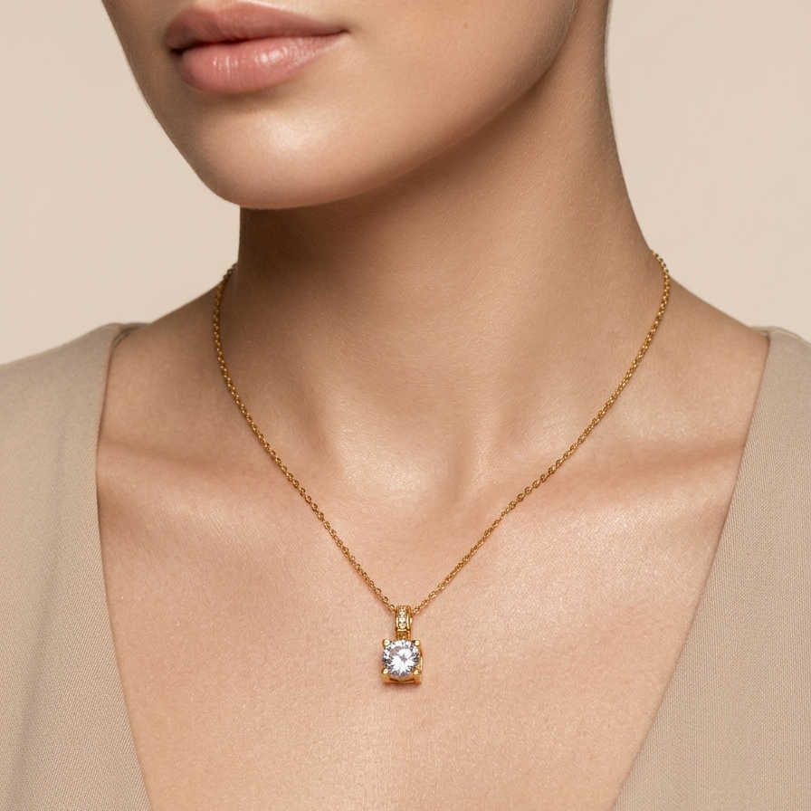 A close-up of a model wearing a delicate gold chain necklace with a single bezel-set clear gemstone pendant, styled with a beige top against a soft beige background.