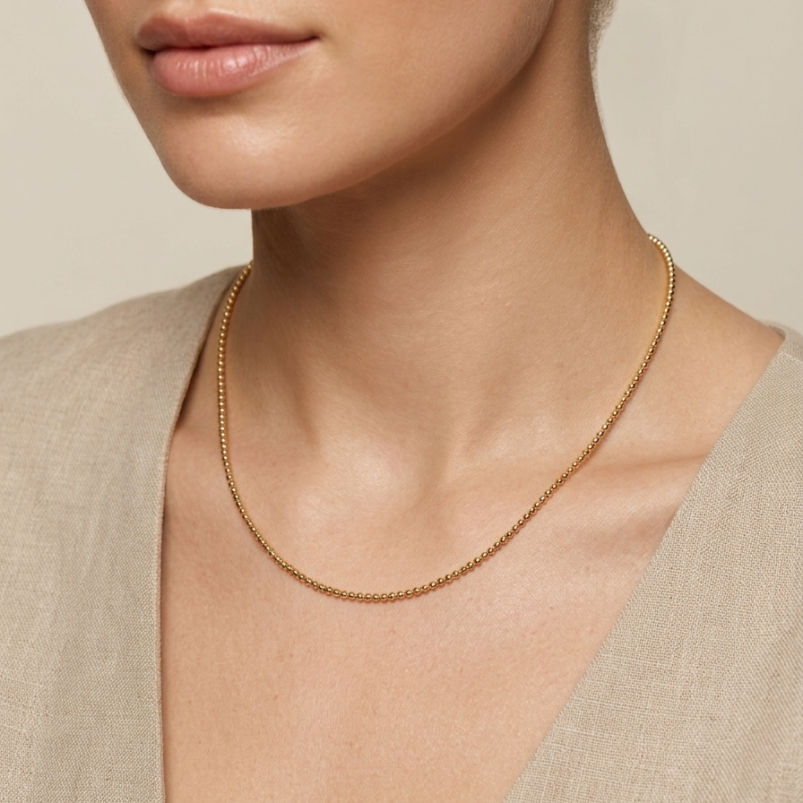 A close-up of a model wearing a delicate gold chain necklace with a textured, beaded appearance, styled with a beige linen top against a soft beige background.