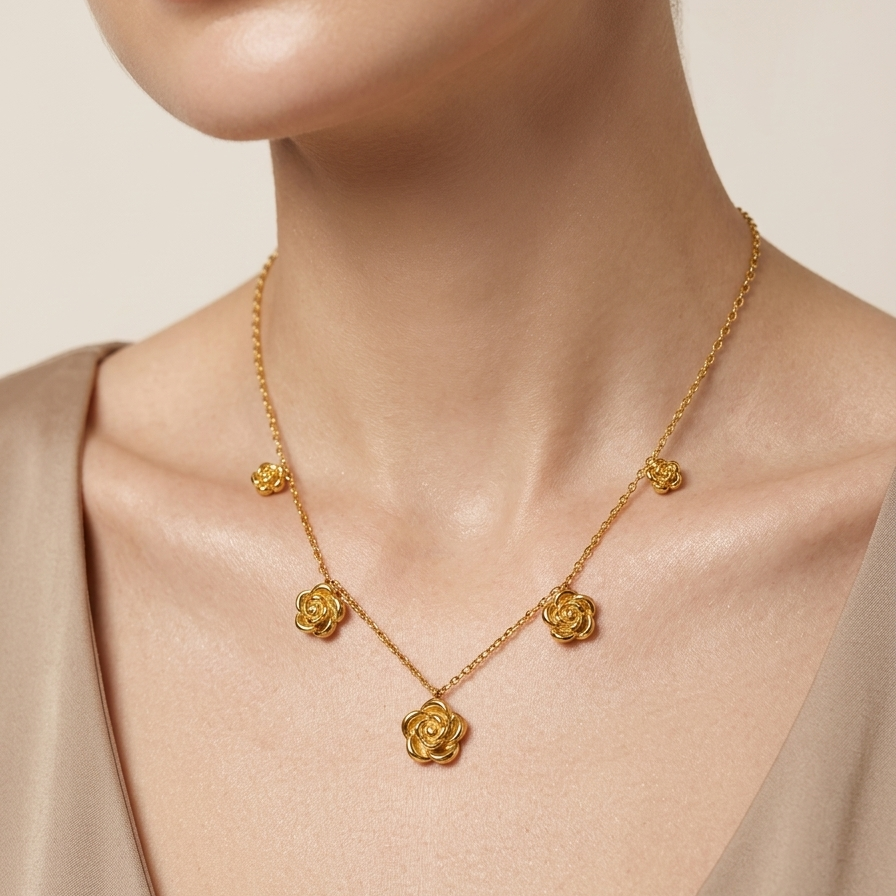 A close-up of a model wearing a delicate gold necklace with five rose-shaped charms spaced along a fine chain, styled with a beige satin top against a soft beige background.