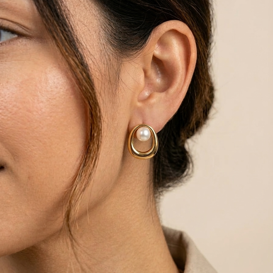 Close-up of a person wearing a 18k gold plated stud earrings featuring an open oval design with a single white pearl.