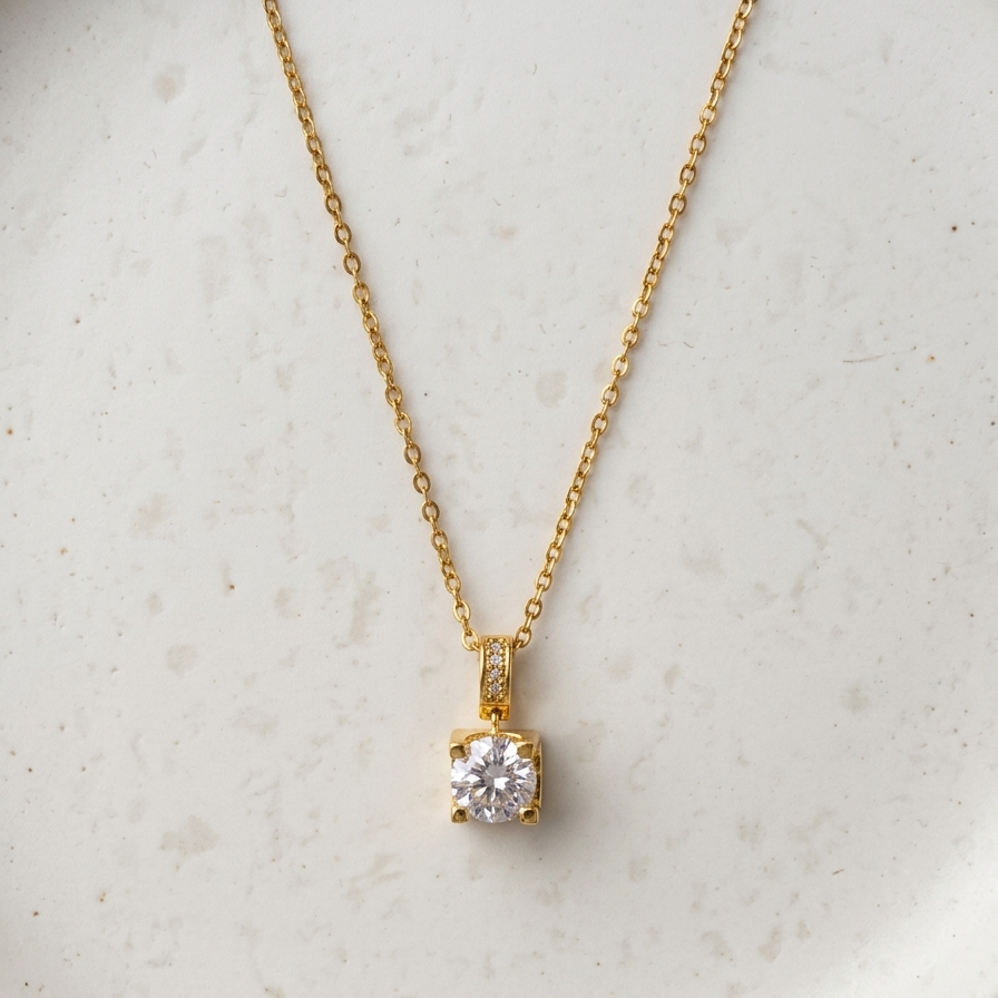 A delicate gold chain necklace with a small round-cut clear gemstone pendant, displayed on a textured white ceramic plate.