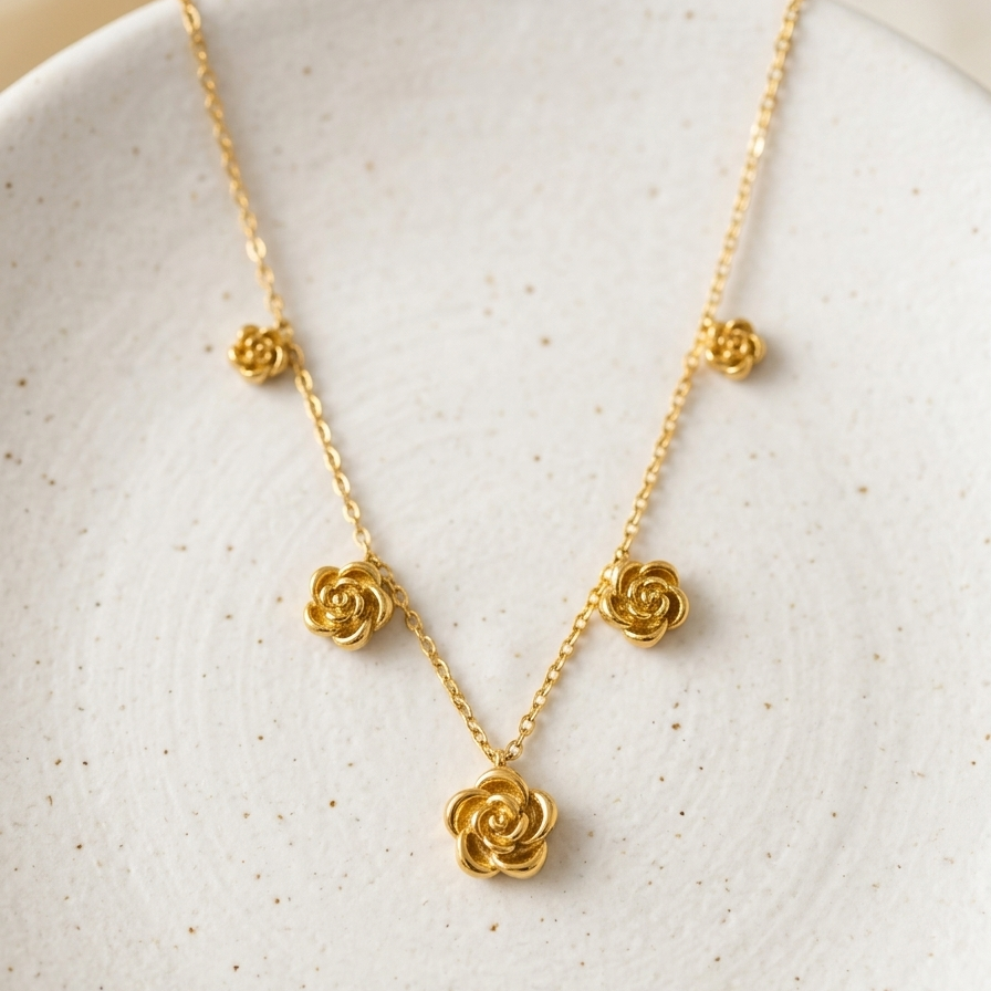 A delicate gold necklace with five small rose-shaped charms and one larger central rose pendant, displayed on a speckled off-white ceramic plate.