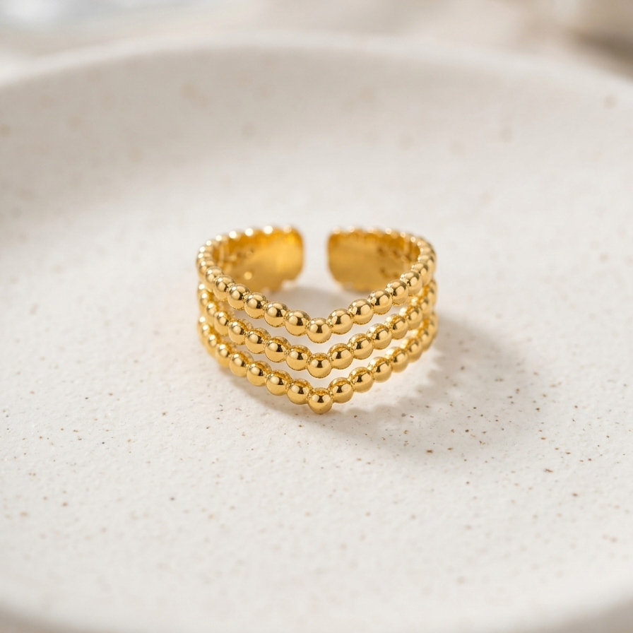 A gold-toned three-band beaded ring displayed on a textured white ceramic dish.