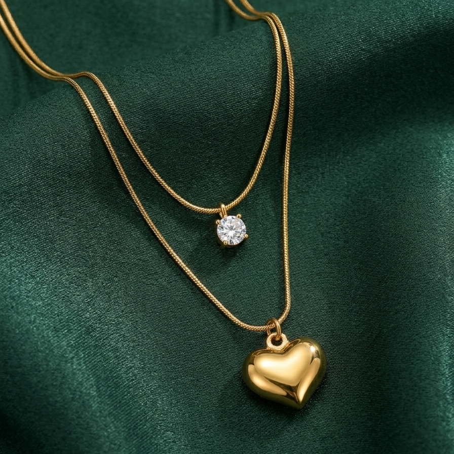 Double layered 18kt gold plated necklace with crystal solitaire and puffy heart pendant on green fabric background.