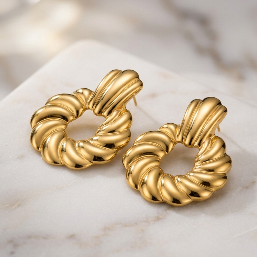 A pair of chunky gold-toned twisted hoop earrings with a ribbed, rope-like texture, displayed on a white marble surface.