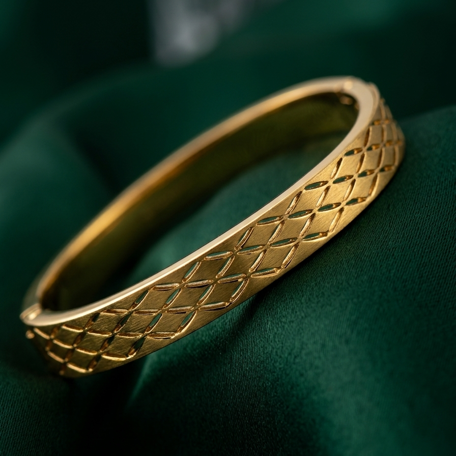 A gold bangle bracelet with a textured diamond-patterned surface, resting on a dark green satin fabric background.