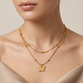 A close-up of a model wearing a delicate double-layered gold necklace with a small butterfly pendant, styled with a beige satin top against a soft beige background.
