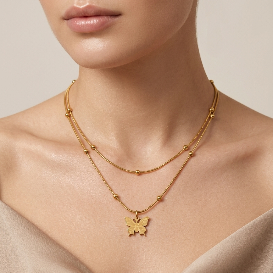 A close-up of a model wearing a delicate double-layered gold necklace with a small butterfly pendant, styled with a beige satin top against a soft beige background.