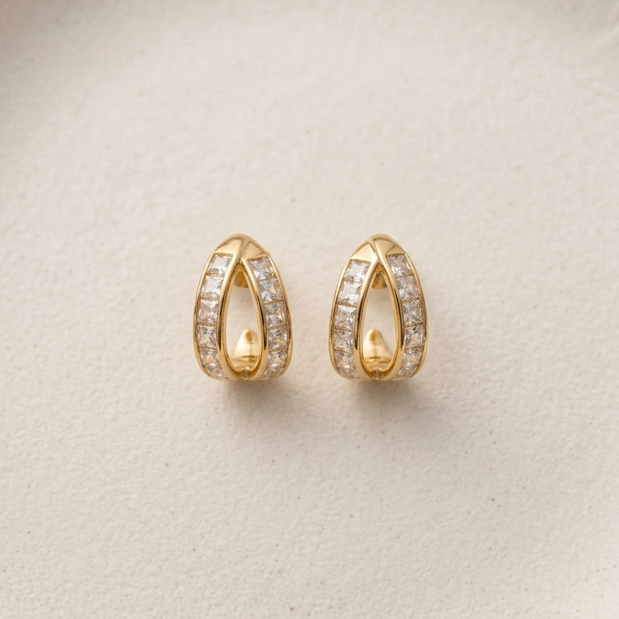 A pair of gold huggie hoop earrings with pavé-set clear stones, displayed on a textured beige surface.