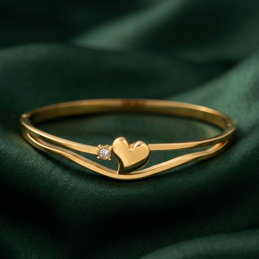 A gold-plated double-band bangle bracelet with a small heart charm and a tiny clear gemstone, displayed on a dark green satin fabric background.