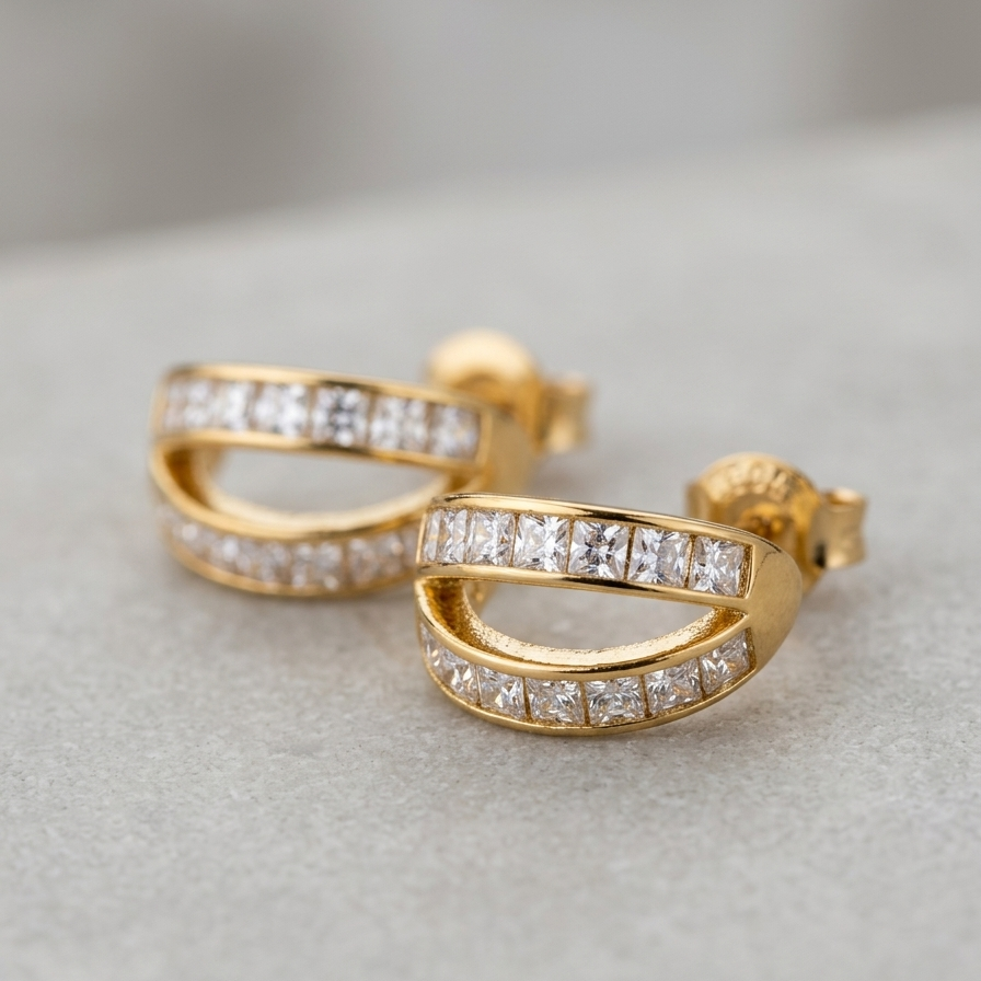 A pair of gold hoop-style earrings with rows of clear square-cut stones, photographed on a soft light gray surface.