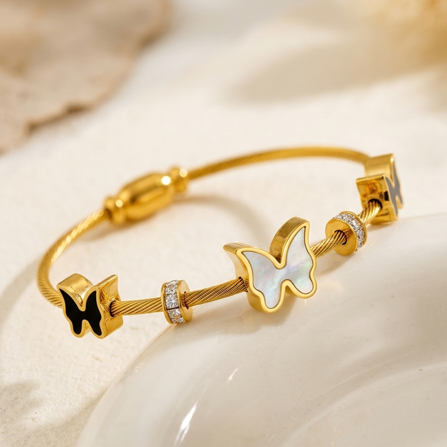 A close-up photography of 18kt gold plated cable wire magnetic bangle bracelet with black and white butterfly charms on white plate.