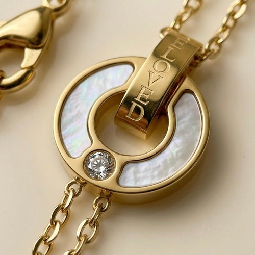 A close-up of 18kt gold plated, waterproof chain bracelet with interlocking circles, mother of pearl inlay and engraved "BELOVE" text on biege background.