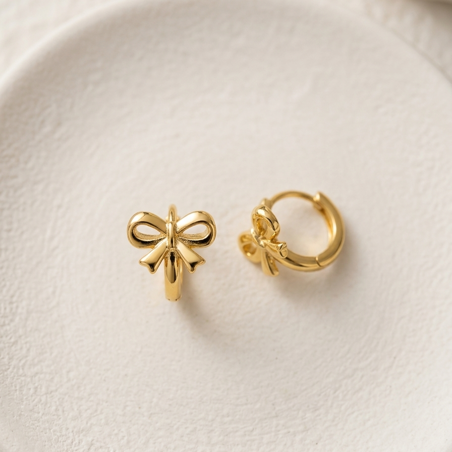 18kt gold plated ribbon bow huggies on white ceramic plate.