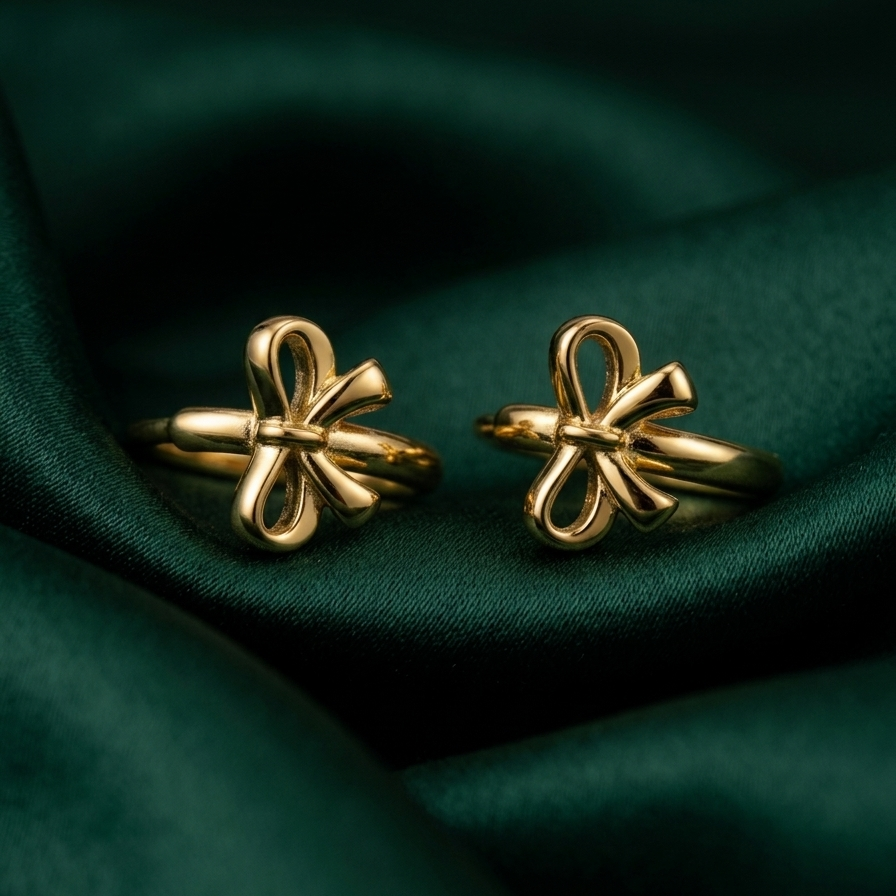 18kt gold plated ribbon bow huggies resting on a green fabric .