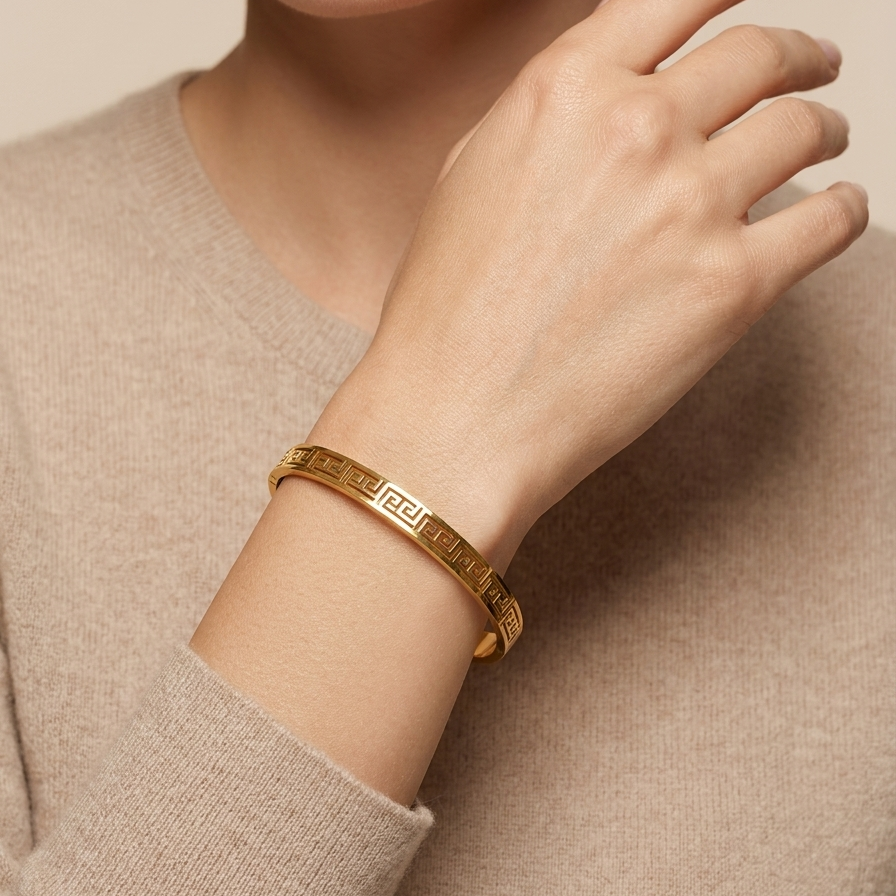 A woman model worn 18kt gold plated, waterproof oval bangle bracelet with cut-out Greek Key pattern on her wrist.