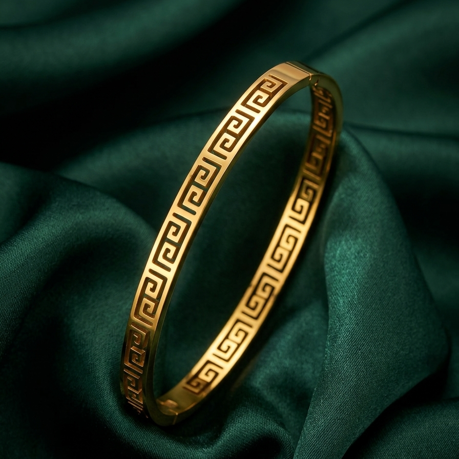 18kt gold plated greek key bangle bacelet resting on smooth green fabric.