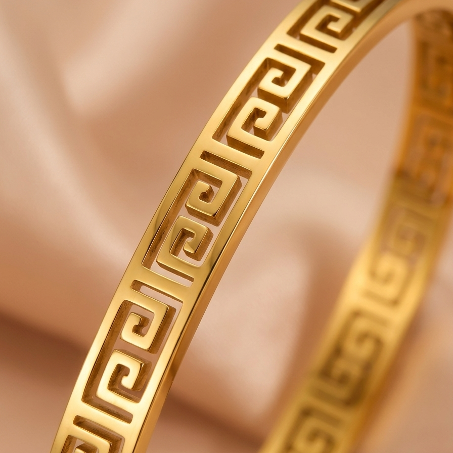 A close-up of 18kt gold plated, waterproof oval bangle bracelet with cut-out Greek Key pattern on beige background.