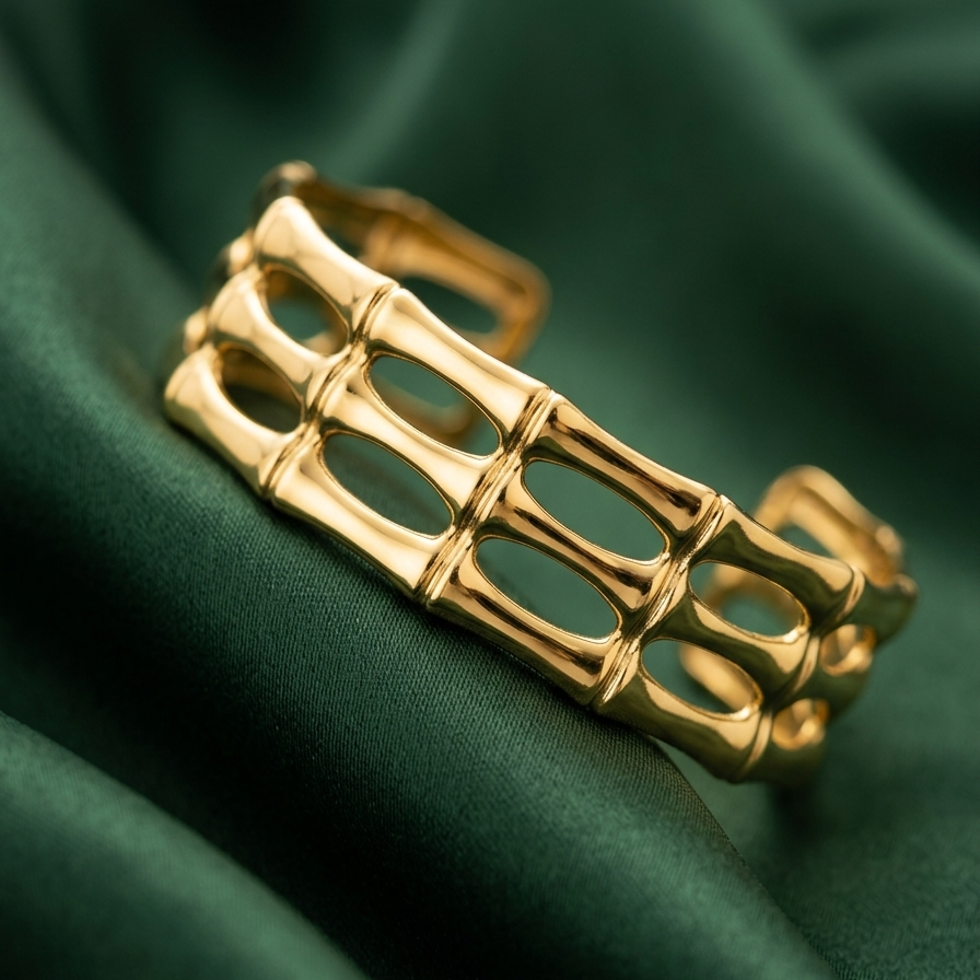 18kt wide gold plated, waterproof cuff bracelet with openwork bamboo design resting on smooth green fabric.