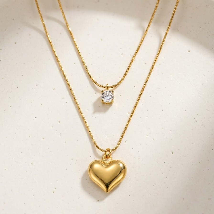 Double layered 18kt gold plated necklace with crystal solitaire and puffy heart pendant on off-white plate.