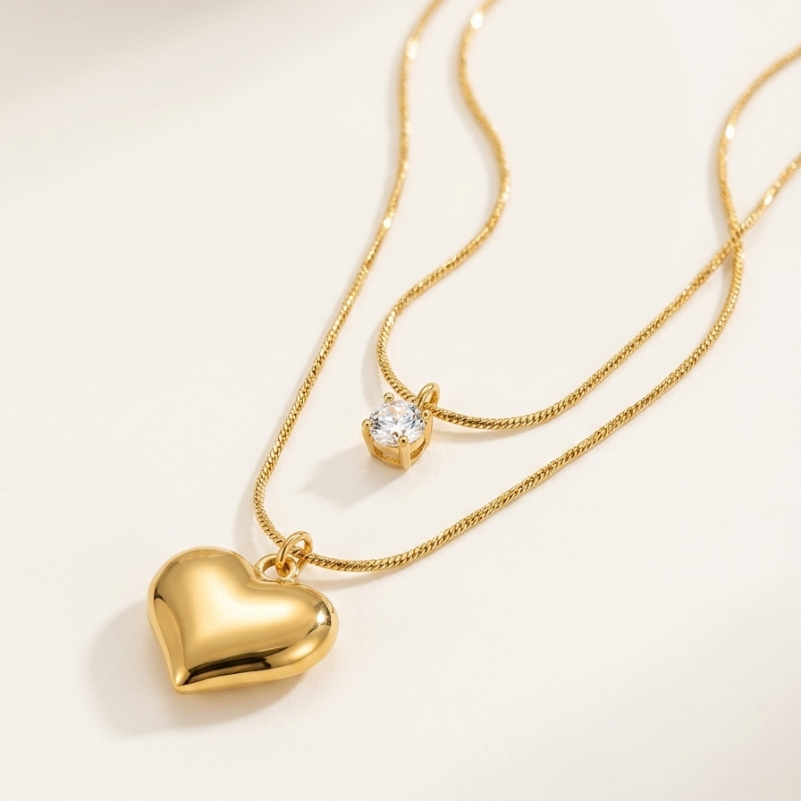 A close-up of double layered 18kt gold plated necklace with crystal solitaire and puffy heart pendant on beige background.
