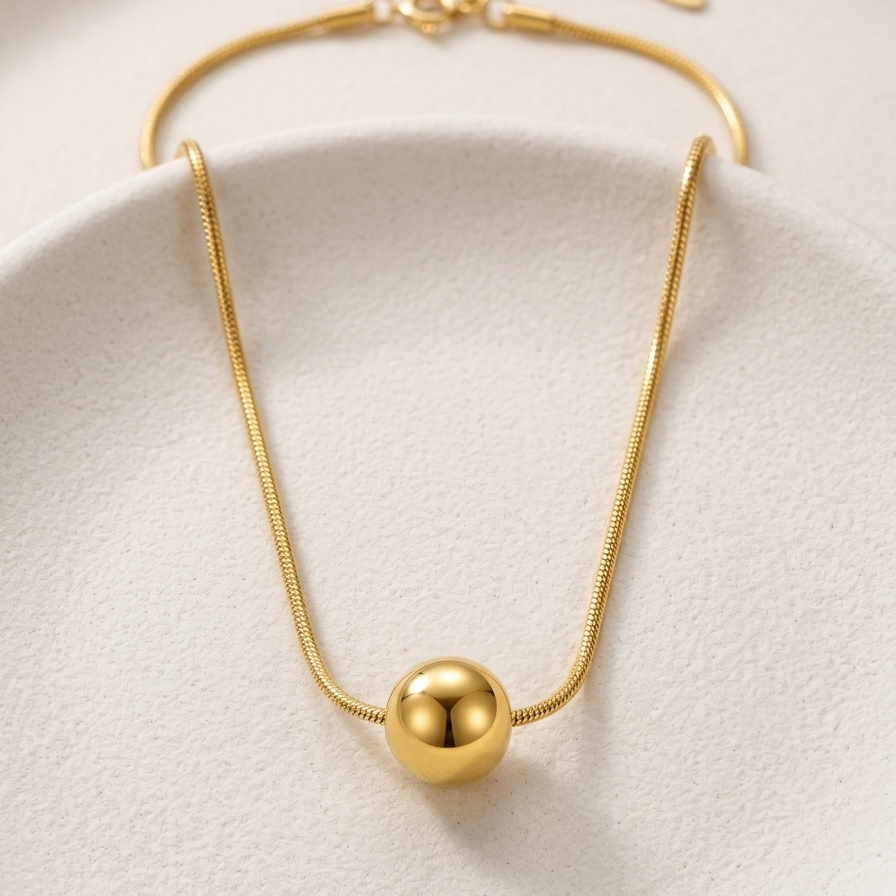 A gold snake chain necklace with a polished spherical bead pendant, displayed on a textured off-white ceramic plate.