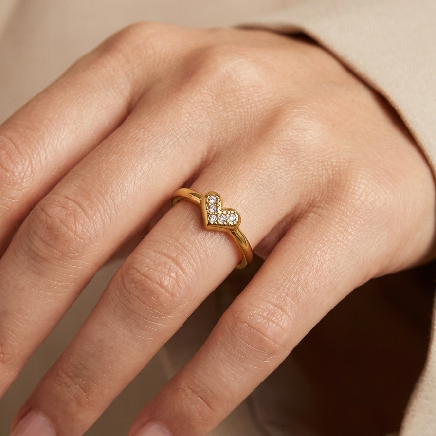 A gold ring with a small heart-shaped pavé setting worn on a person's ring finger, shown in a close-up shot against a soft beige background.