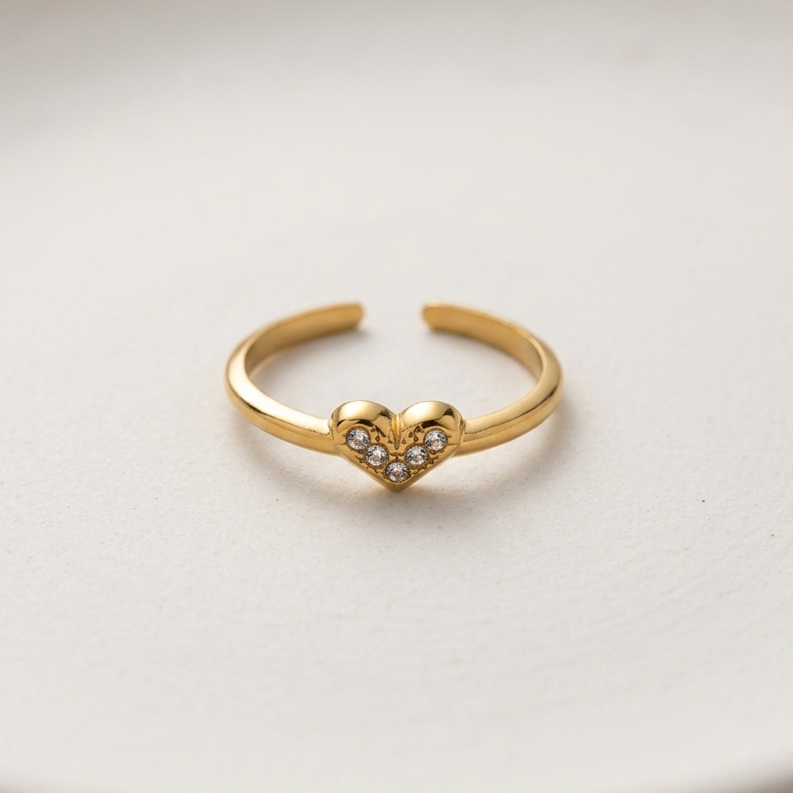 A gold adjustable ring with a small heart-shaped pavé center, resting on a light beige textured surface.
