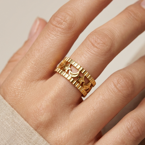 A close-up of a hand wearing a wide gold ring with a textured, ridged outer band and a smooth inner band featuring crescent moon and star motifs.
