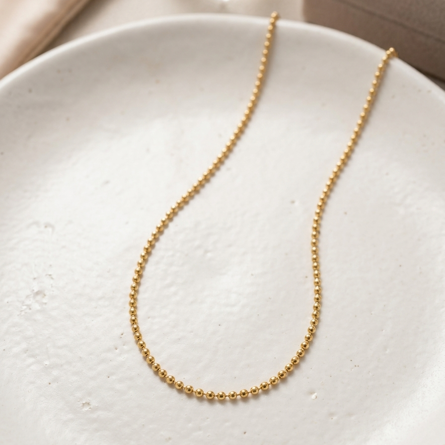 A delicate gold beaded chain necklace draped in an oval shape on a textured white ceramic plate, with a neutral beige jewelry box and soft fabric in the background.
