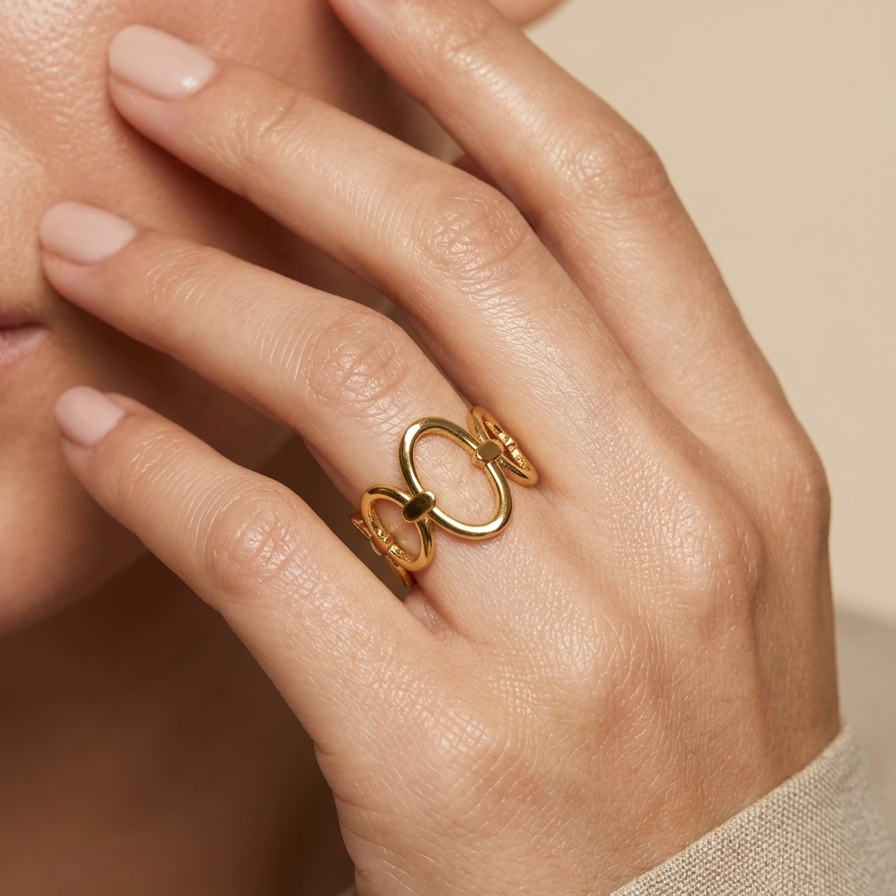 A close-up of a person's hand wearing a gold ring with two interlocking oval shapes, against a soft beige background.