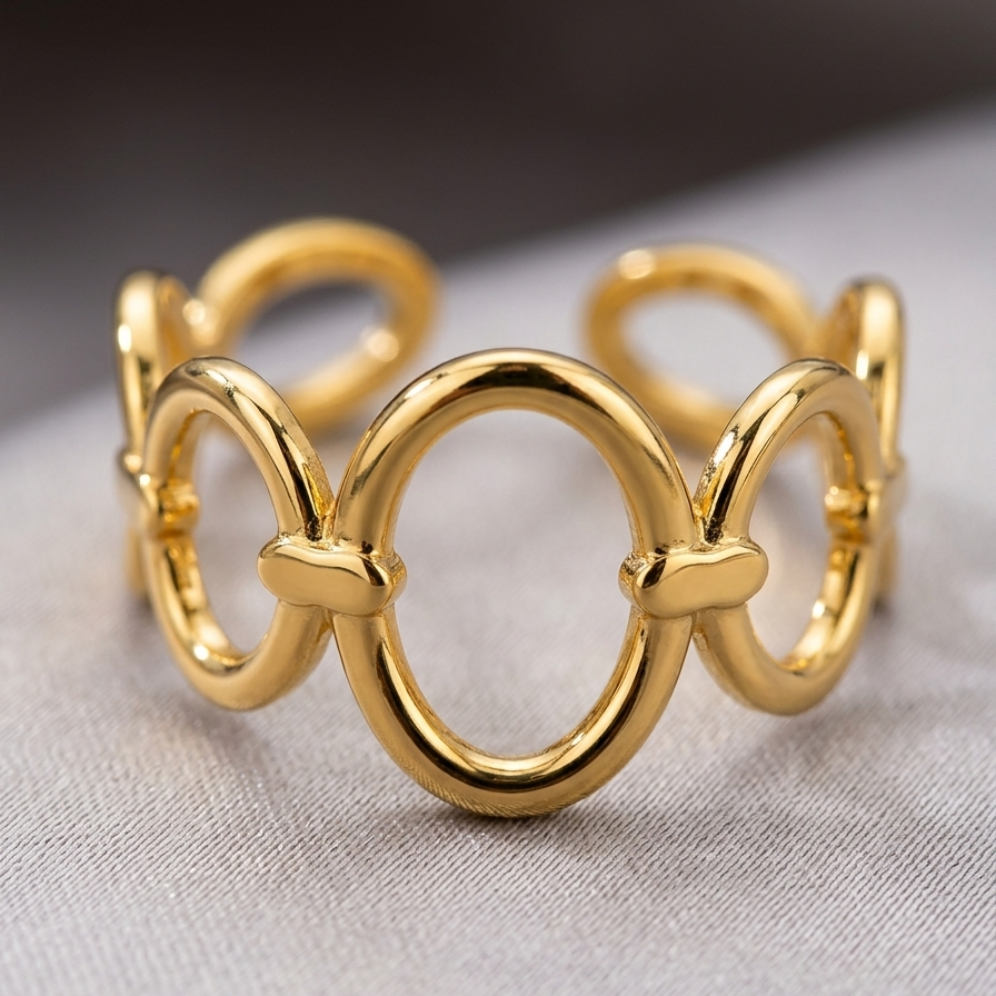 A polished gold-tone metal ring with a continuous interlocking circular and oval link design, resting on a light gray textured surface.