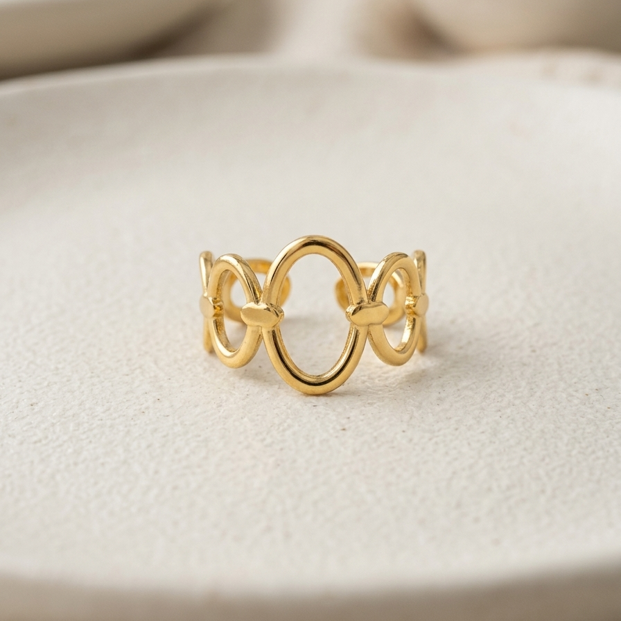 A delicate gold ring with three interlocking oval loops, resting on a textured beige ceramic plate with a soft-focus background.