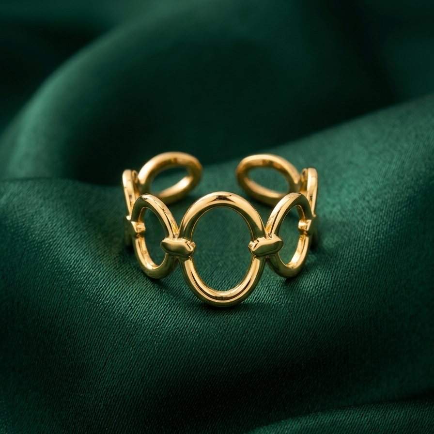 A gold-toned open ring band with interlocking circular and looped details, displayed on draped dark green satin fabric.