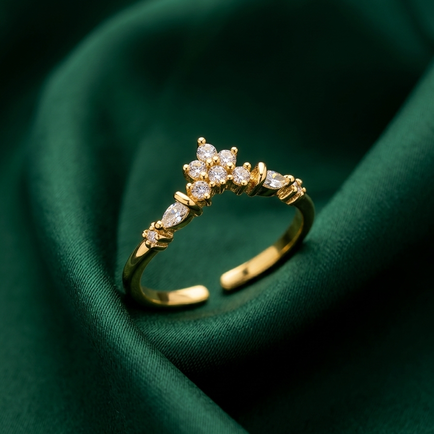 A gold adjustable ring with a floral cluster of clear stones, resting on draped dark green satin fabric.