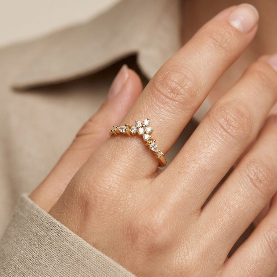 A close-up of a hand wearing a delicate gold ring with small clear stones arranged in a floral or leaf-like cluster, against a soft beige background.