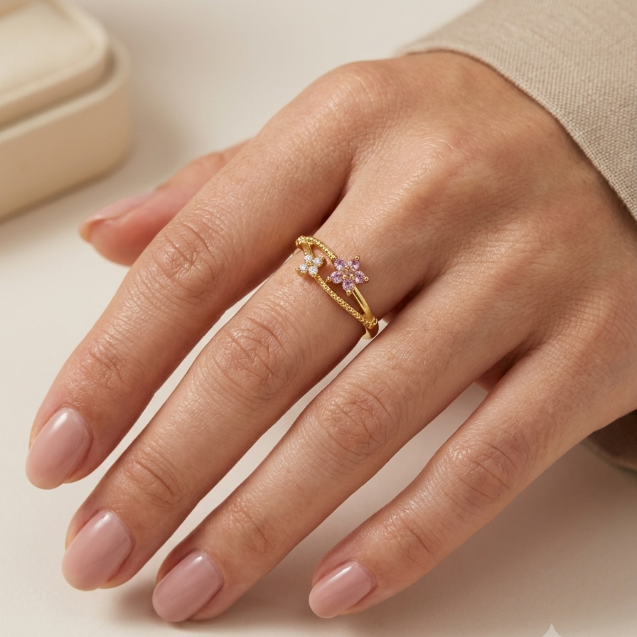 A close-up of a hand wearing a delicate gold ring with a small pink gemstone and a clear accent stone, set against a soft beige background.