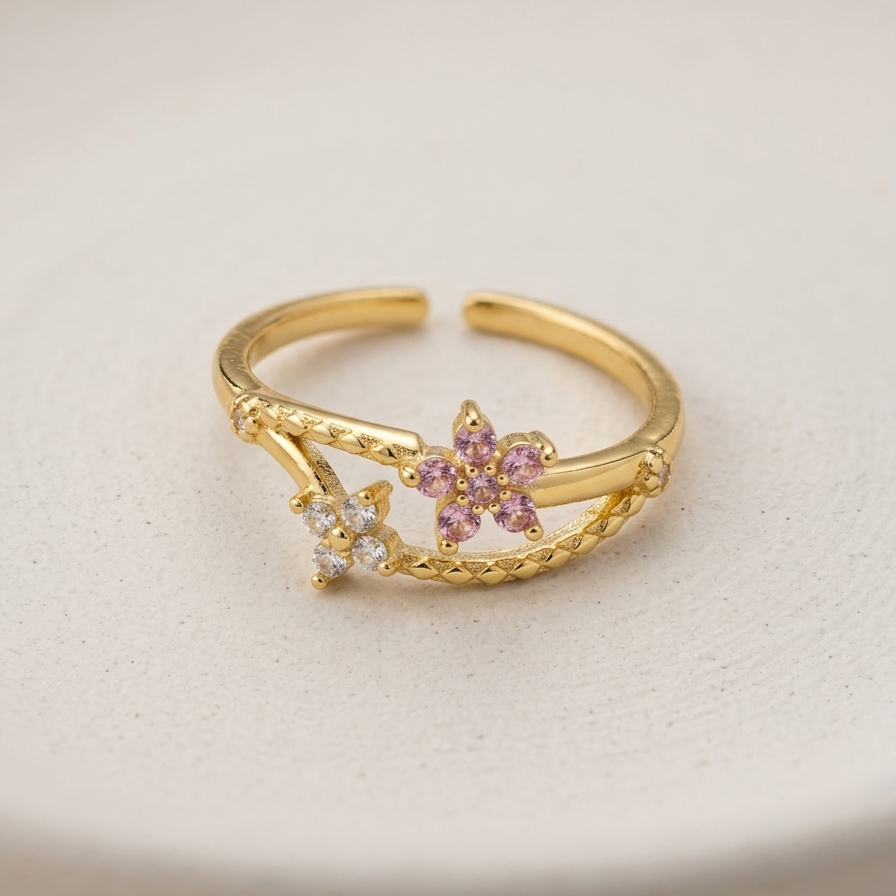 A gold-plated adjustable ring with two flower motifs, one in light pink stones and one in clear stones, resting on a textured off-white surface.