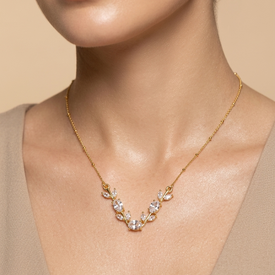 A close-up of a model wearing a delicate gold necklace with a V-shaped floral cluster of clear crystals, set against a soft beige background.