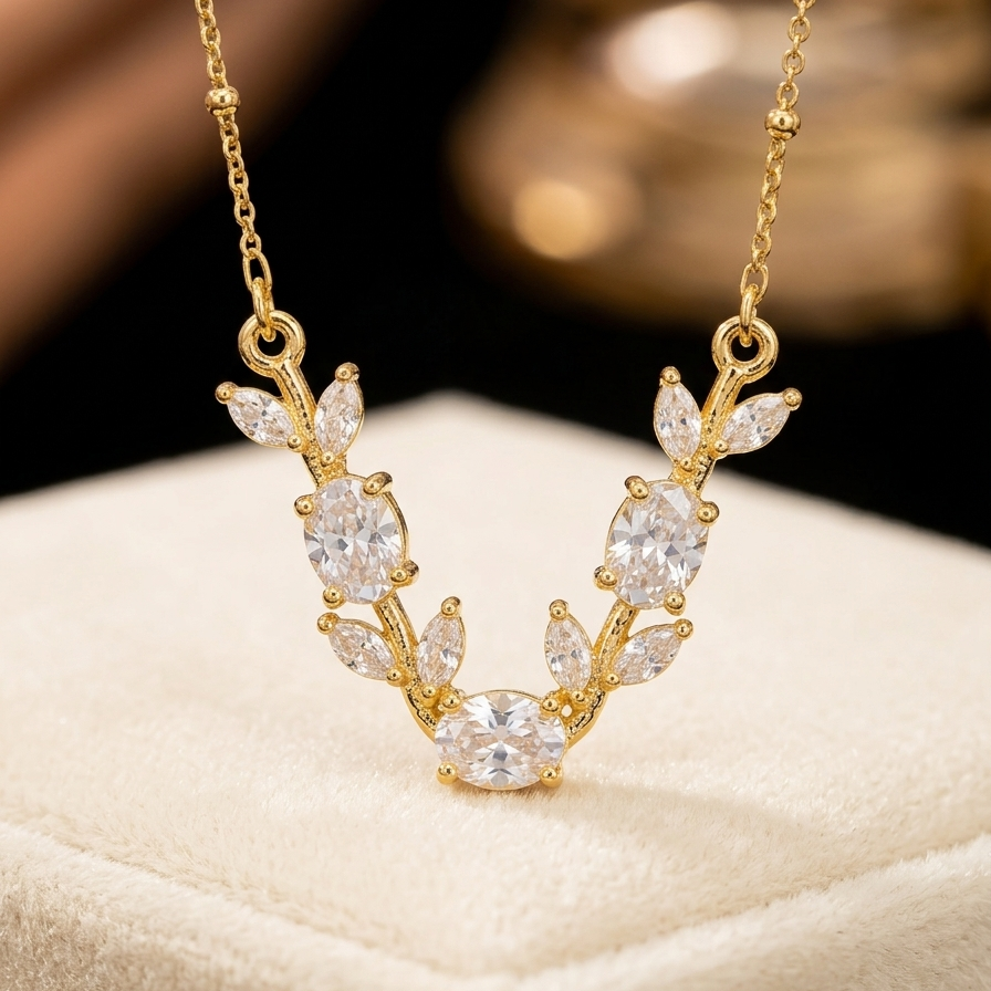 Gold leaf-shaped pendant necklace with clear gemstones, displayed on a cream jewelry box against a dark blurred background.