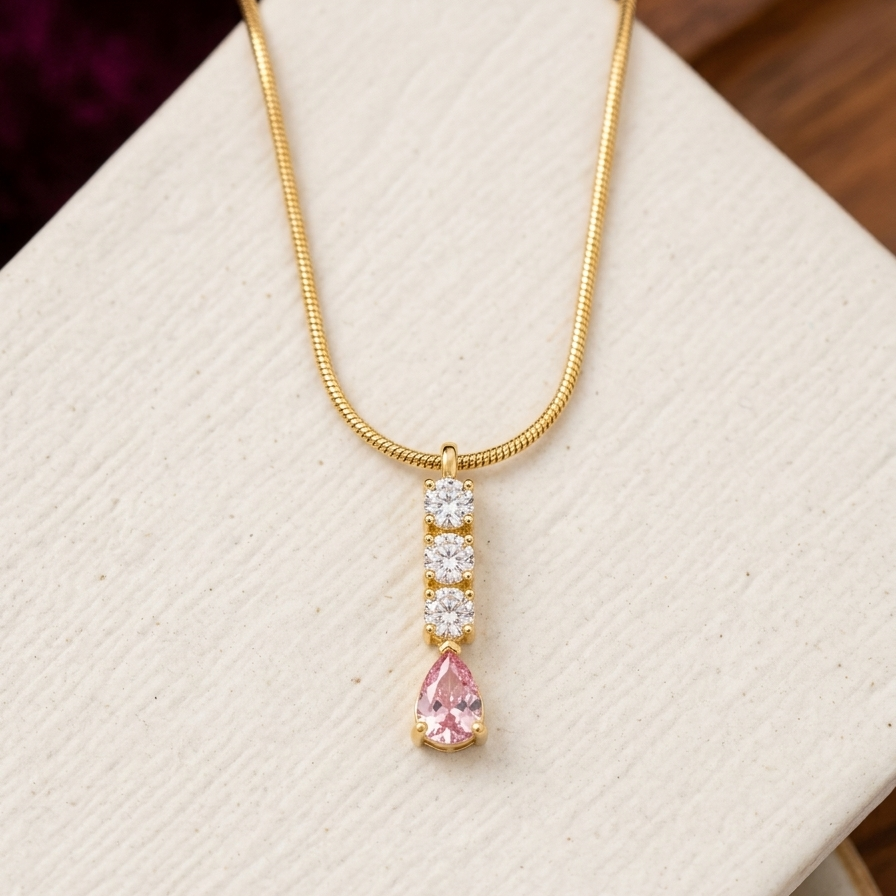 A gold snake chain necklace with a vertical pendant featuring three clear round-cut stones and a teardrop-shaped pink gemstone at the bottom, displayed on a textured cream jewelry box.