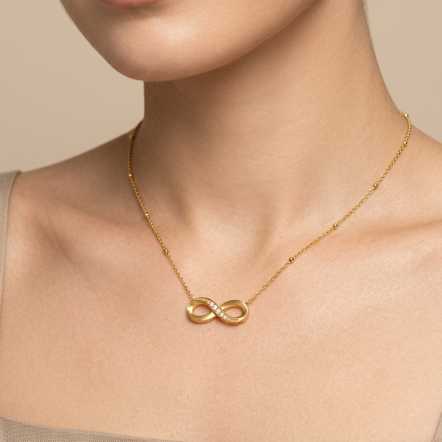 A close-up of a model wearing a delicate gold infinity necklace with a polished finish, styled with a beige linen top against a soft beige background.