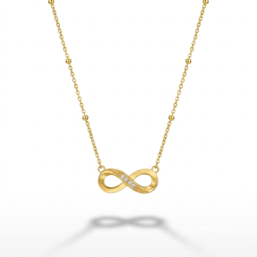 A delicate gold chain necklace featuring an infinity symbol pendant with a row of small clear stones, displayed on a clean white background with a soft reflection below.
