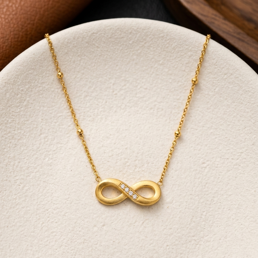 A gold infinity necklace with a small row of clear stones across the center, displayed on a textured white jewelry stand.