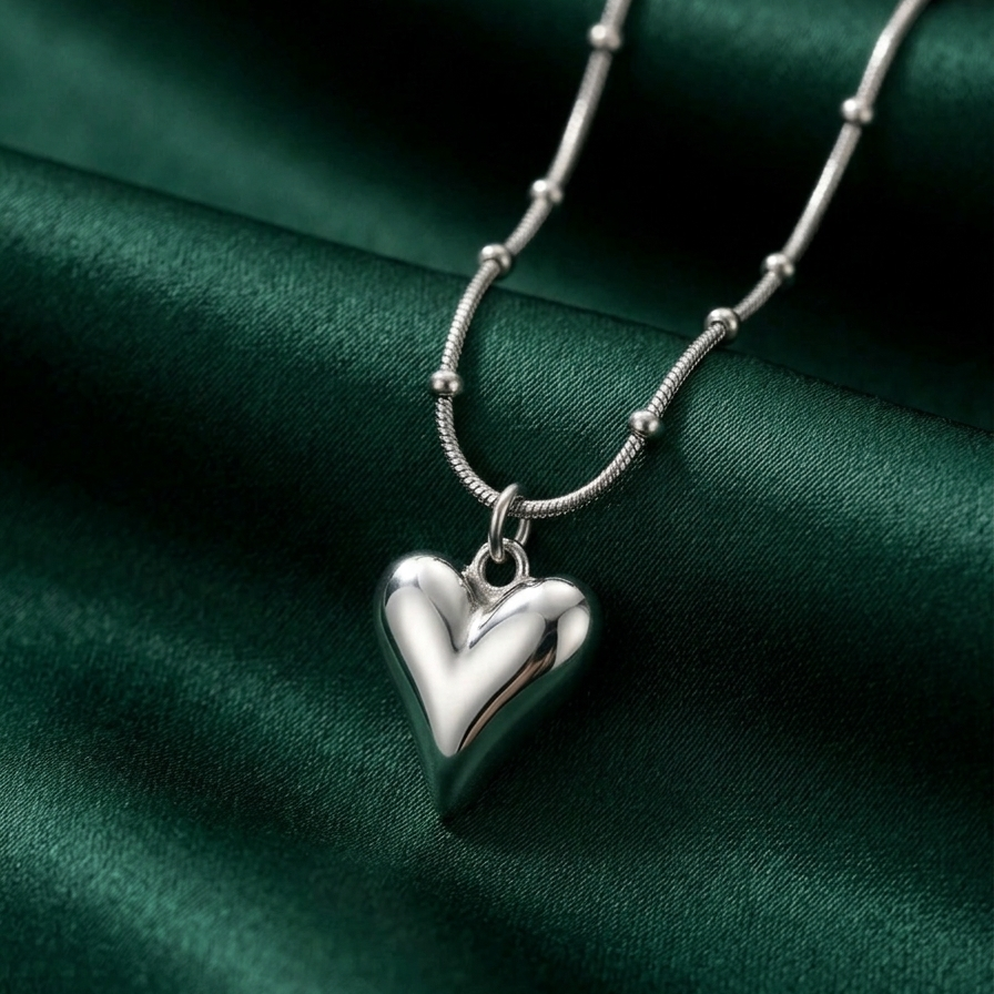 Silver-tone rhodium plated necklace featuring a smooth, 3D puffy heart pendant suspended on a delicate beaded satellite chain against a green satin background