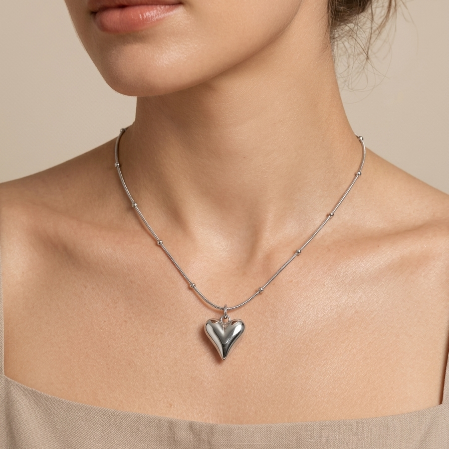 Silver-tone rhodium plated necklace featuring a smooth, 3D puffy heart pendant suspended on a delicate beaded satellite chain  worn by a model on a beige background