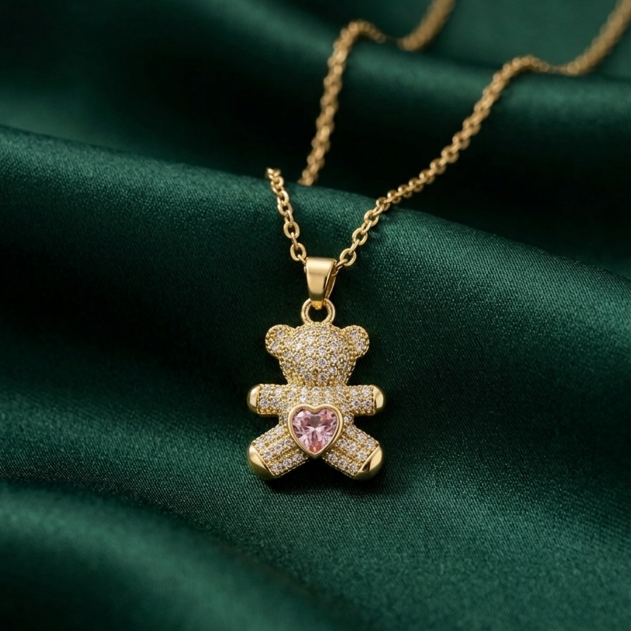 18k gold plated teddy bear pendant necklace fully encrusted with clear CZ diamonds, featuring a pink heart-shaped crystal in the center on a green fabric background