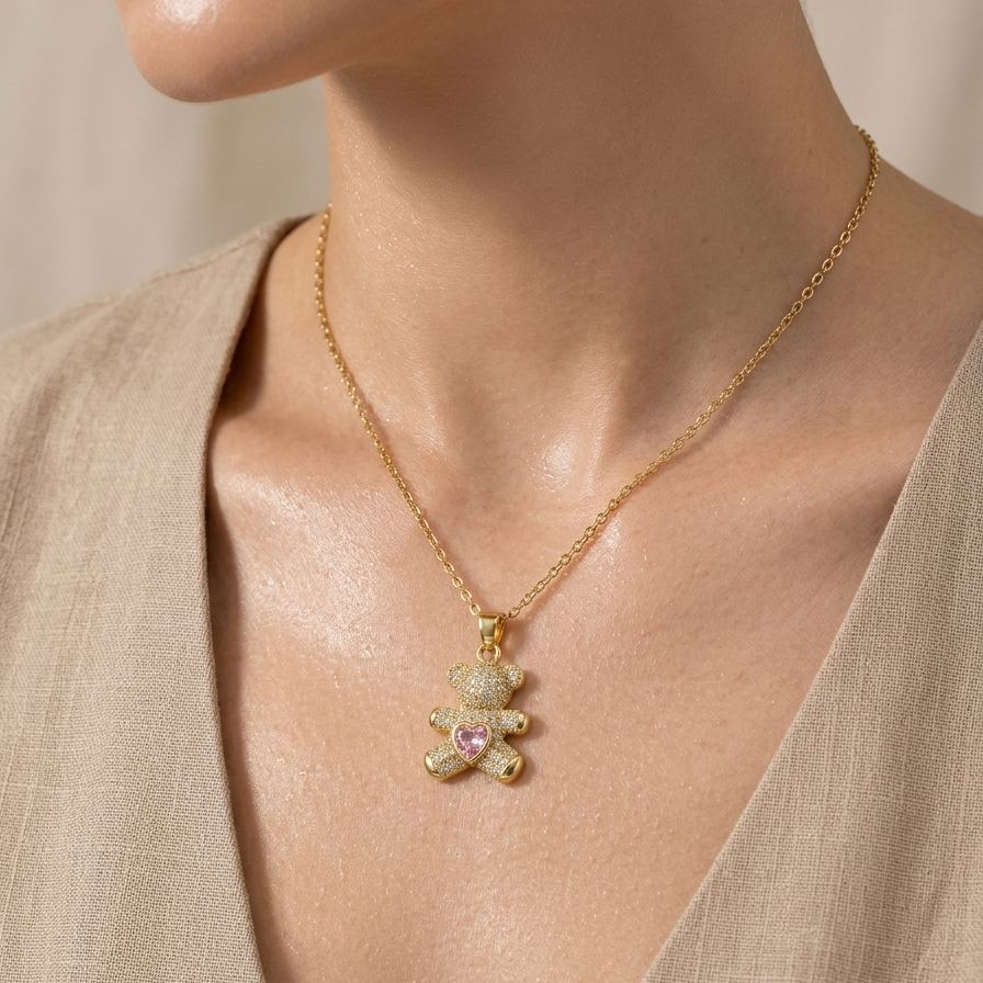18k gold plated teddy bear pendant necklace fully encrusted with clear CZ diamonds, featuring a pink heart-shaped crystal in the center worn by a person in a beige outfit.