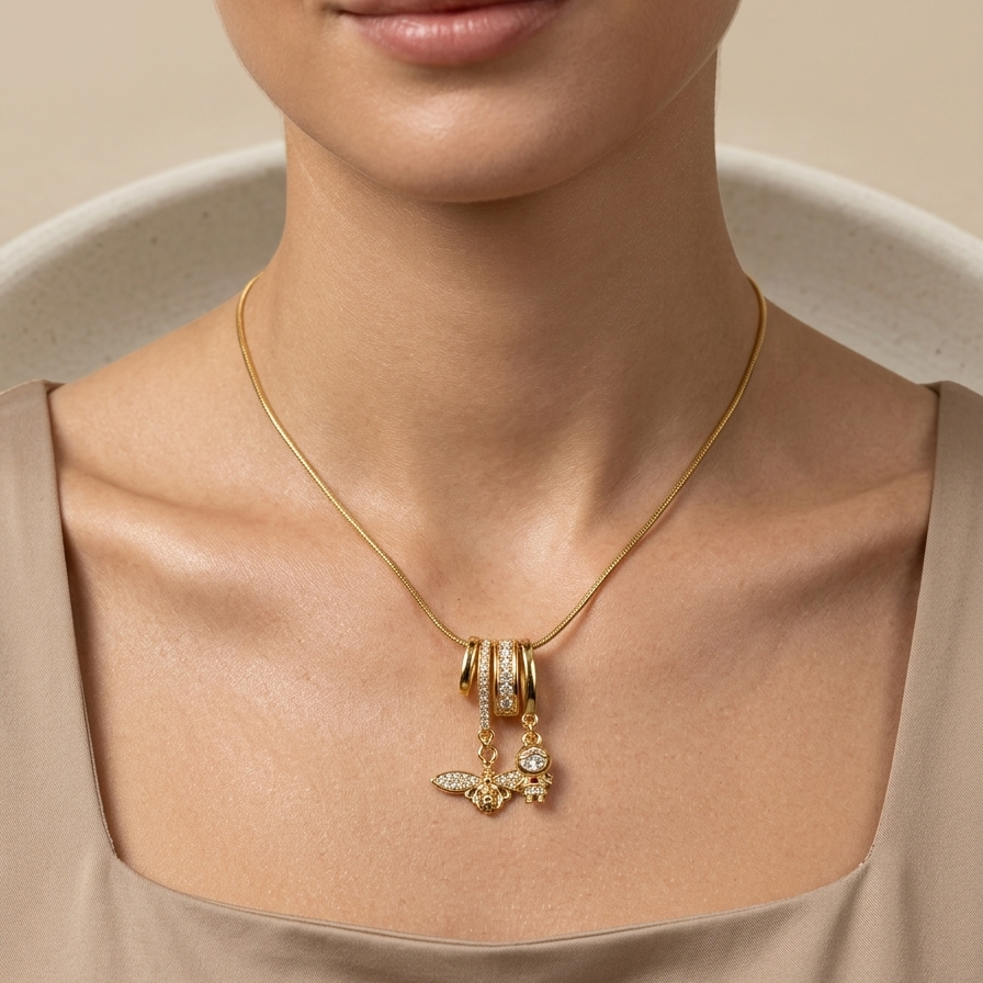 Close-up of a woman wearing 18k gold plated necklace featuring an intricate diamond-encrusted sliding bail holding a detailed golden bee charm and a small figure charm on a beige background