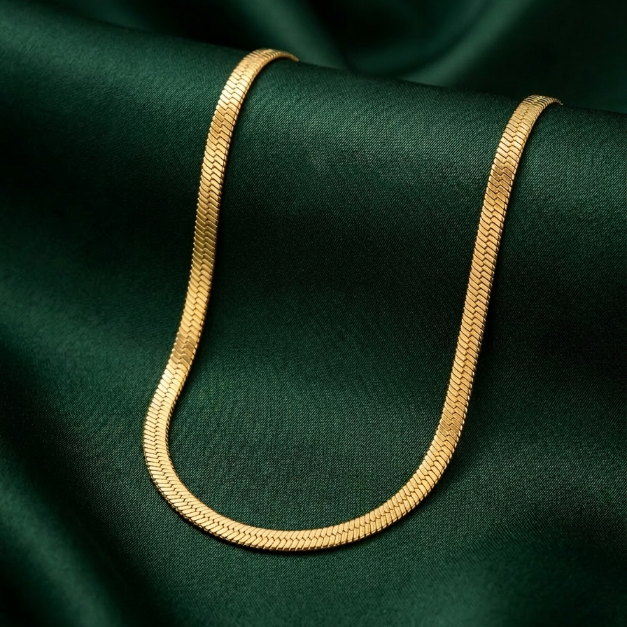 18k gold plated thick, flat herringbone chain necklace resting on a textured white surface on a green fabric background